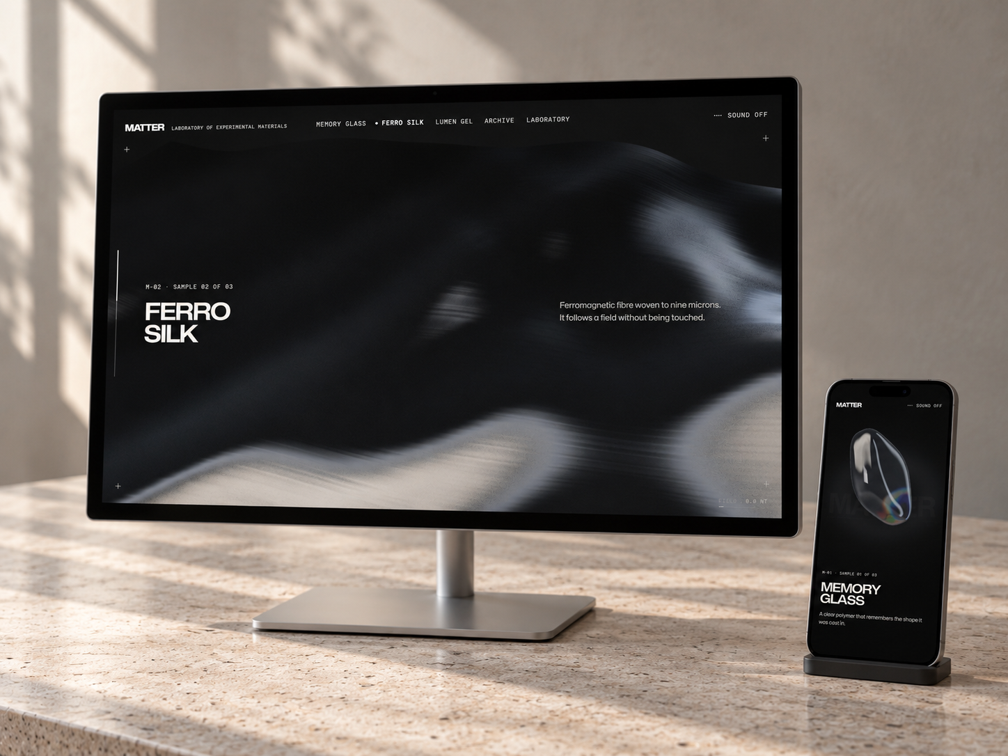 Desktop monitor and smartphone displaying the MATTER website by Fieldmoth on a light stone desk. The monitor shows the dark, flowing texture of Ferro Silk; the phone shows the transparent Memory Glass